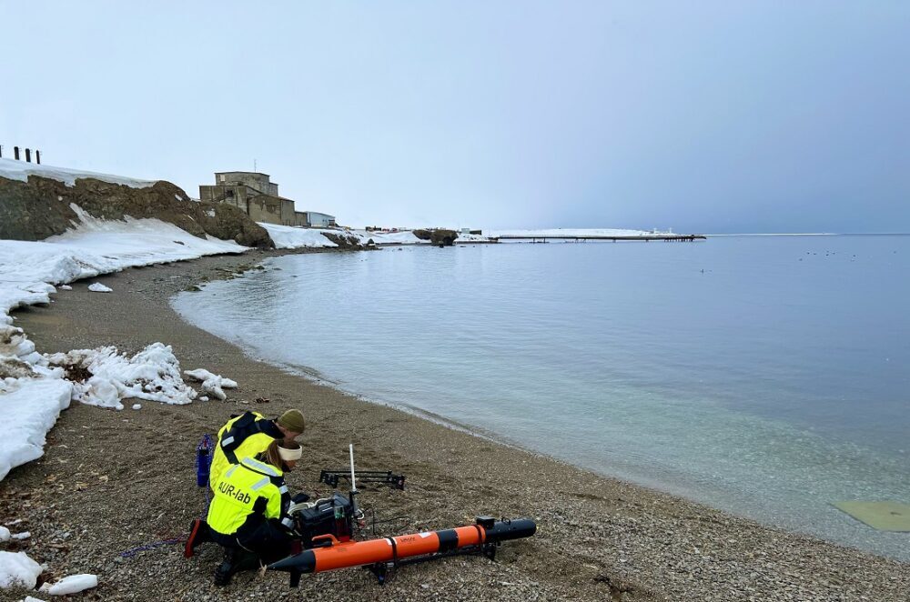 Bilde av to personer med en robot i strandkanten. Artikkelen handler om autonome robotorganisasjoner.