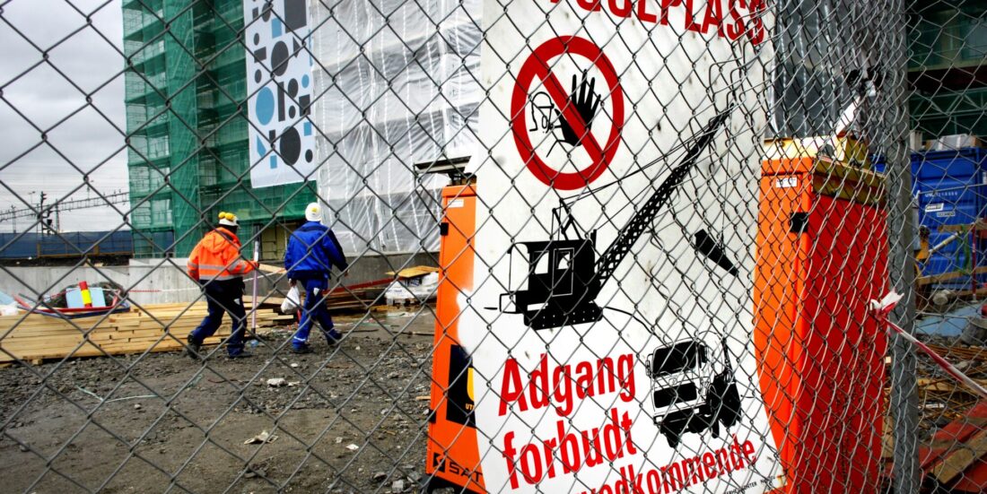 Bilde av byggeplass. Høybygg i bakgrunnen, med to bygningsarbeidere foran. I forgrunnen dominrer et nettinggjerde. På deet sitter en stor plakat med påskriften "Byggeplass - adgang forbudt for uvedkommende"