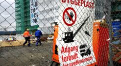 Bilde av byggeplass. Høybygg i bakgrunnen, med to bygningsarbeidere foran. I forgrunnen dominrer et nettinggjerde. På deet sitter en stor plakat med påskriften "Byggeplass - adgang forbudt for uvedkommende"