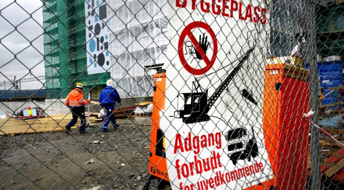 Bilde av byggeplass. Høybygg i bakgrunnen, med to bygningsarbeidere foran. I forgrunnen dominrer et nettinggjerde. På deet sitter en stor plakat med påskriften "Byggeplass - adgang forbudt for uvedkommende"