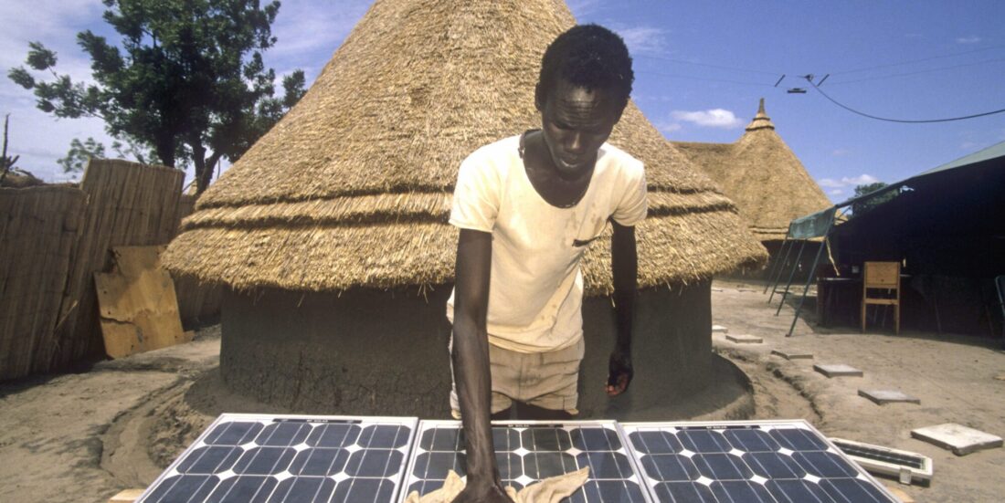 Gutt pusser på solcellepanel foran afrikansk landsbyhus.