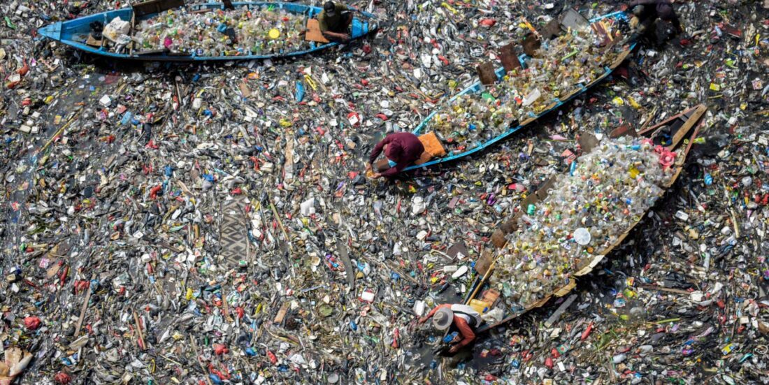Luftfoto av elv som er tettpakket med plastsøppel. Vi ser folk i tre åpne båter plukke plast.