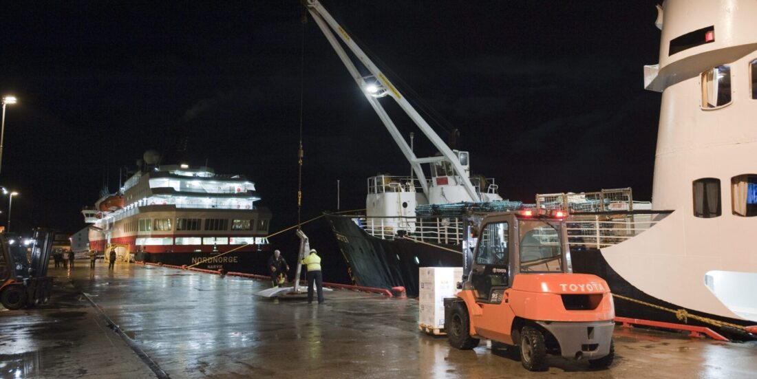 Oransje gaffeltruck kjører langs kai, der to hurtigruteskip ligger i kveldsmørket.