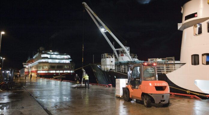 Oransje gaffeltruck kjører langs kai, der to hurtigruteskip ligger i kveldsmørket.
