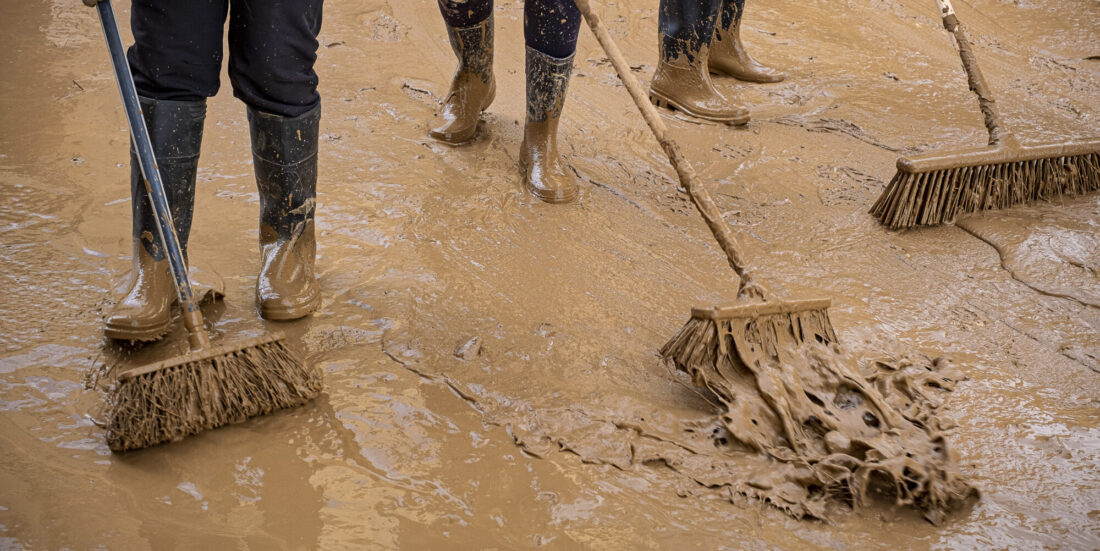 Brun leire ligger i gata, vi ser beina på tre personer i gummistøvler som skyfler leirvannet avgårde med koster