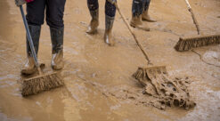 Brun leire ligger i gata, vi ser beina på tre personer i gummistøvler som skyfler leirvannet avgårde med koster