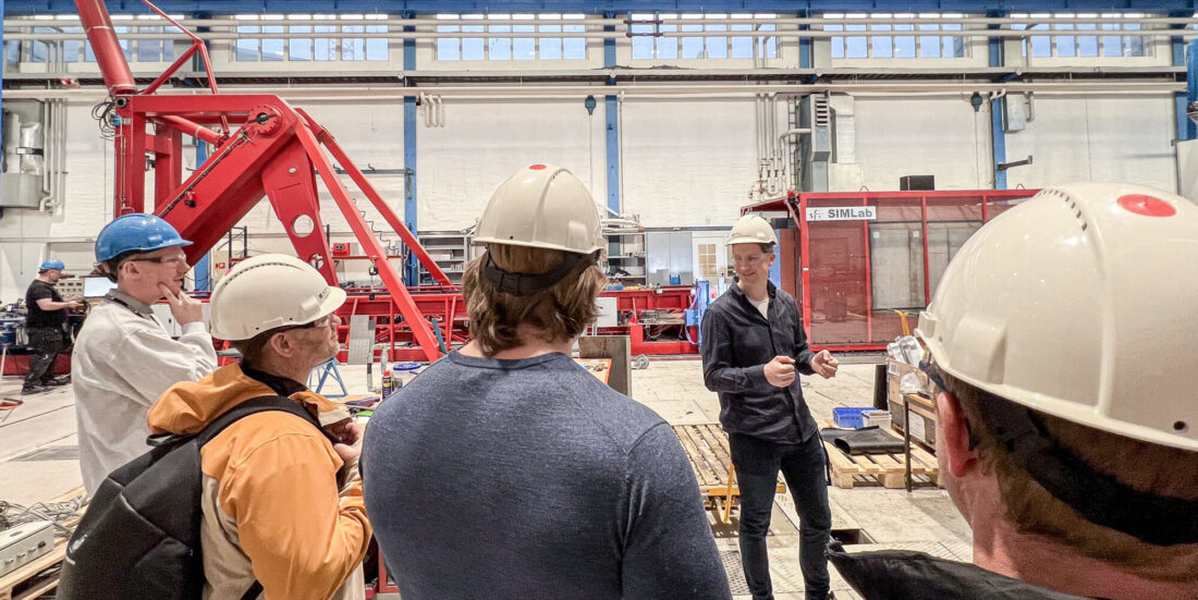 Bilde som viser flere personer med hjelmer i en stor hall med store mekaniske rigger. Bilde til artikkel om testing av rekkverk for broer.