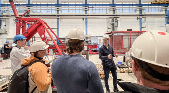 Bilde som viser flere personer med hjelmer i en stor hall med store mekaniske rigger. Bilde til artikkel om testing av rekkverk for broer.