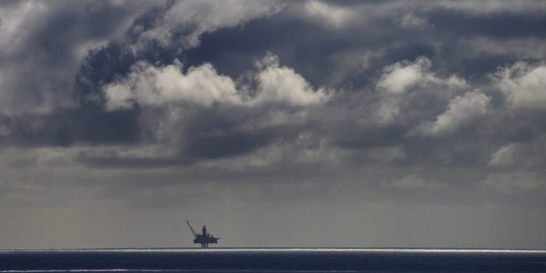 Oljeplattform ses mot horisonten. Solstripe på havet foran plattformen. På himmelen ses dramatiske svarte, grå og hvite skyer.