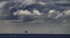 Oljeplattform ses mot horisonten. Solstripe på havet foran plattformen. På himmelen ses dramatiske svarte, grå og hvite skyer.