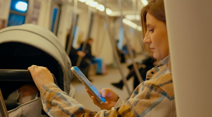 KI. En kvinne på toget scroller på mobilen mens barnevognen står ved siden av henne.