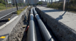 Svarte fjernvarmerør ligger i grøft som er gravd opp. I bakgrunnen skimtes boligblokker.