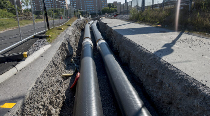 Svarte fjernvarmerør ligger i grøft som er gravd opp. I bakgrunnen skimtes boligblokker.