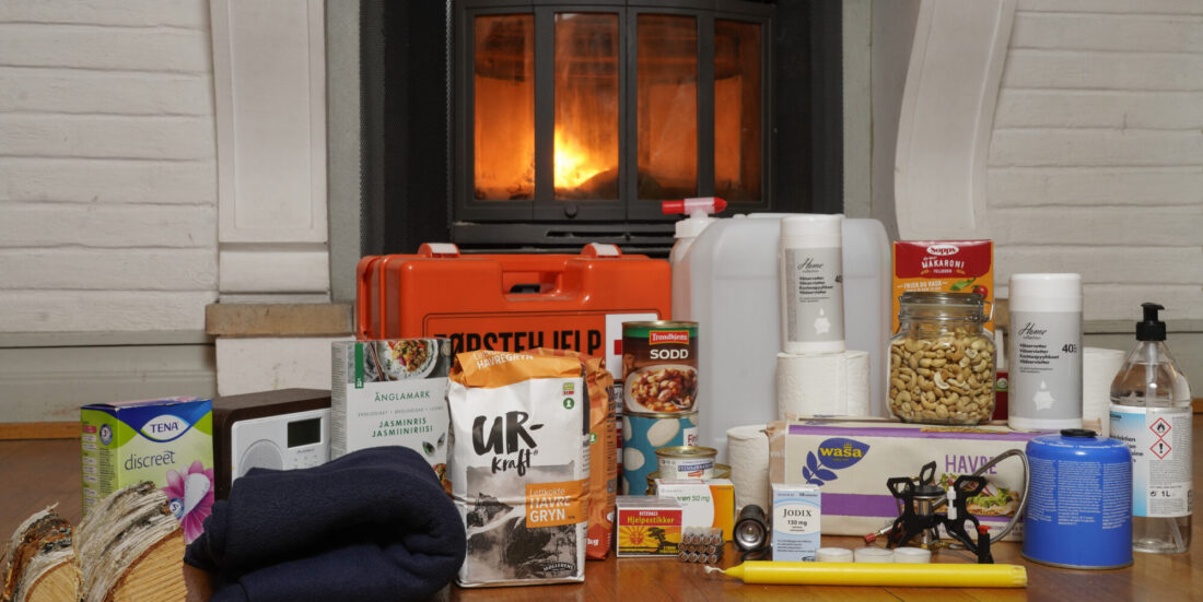 Vannkanner, fyrstikkeksker, primus og mye annet utstyr som inngår i egenberedskap, står på et stuegulv foran en peis som brenner.
