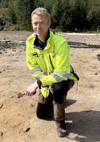 Arkeolog Mjærum sitter på huk og ser mot kamera. Foto.