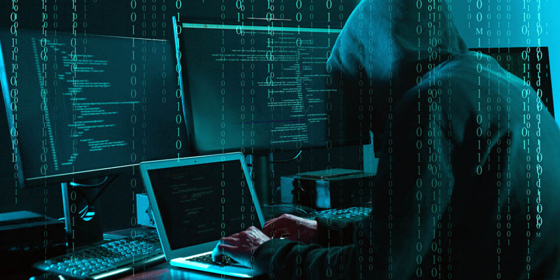 En person med hettegenser sitter foran datamaskiner og ser ut til å gjøre ugagn. Foto.