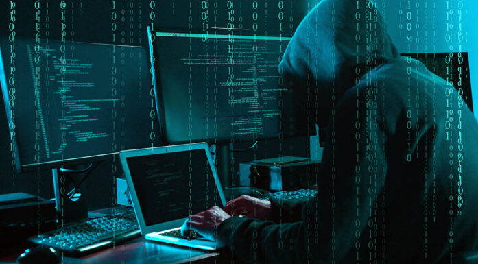 En person med hettegenser sitter foran datamaskiner og ser ut til å gjøre ugagn. Foto.